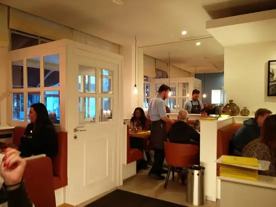 Het interieur van de brasserie met een wit houten tochtportaal, roodbruine zitbanken en gasten aan tafel