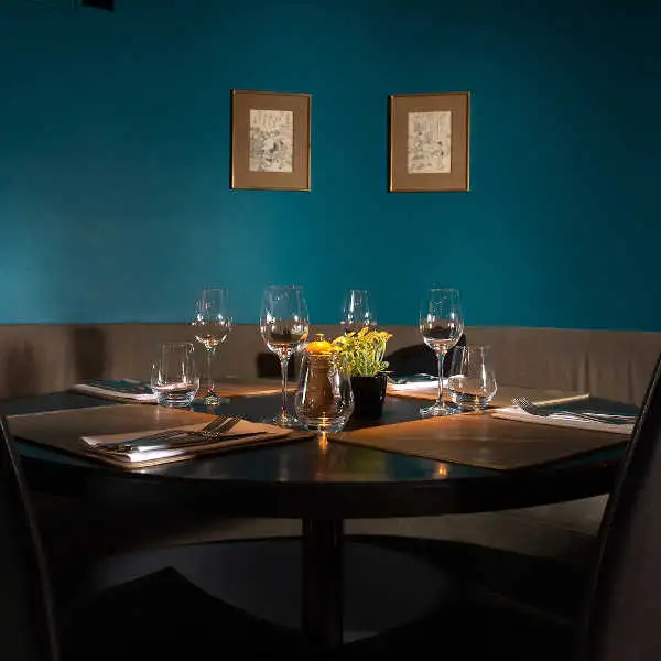 Ronde tafel gedekt met wijnglazen en een kaars, voor een petrolblauwe muur met twee ingelijste prenten