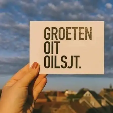 Een hand houdt een kaartje met "Groeten oit Oilsjt" omhoog boven de daken van de stad