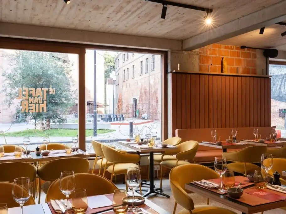 Gedekte tafels met okergele stoelen bij het raam met de naam De Tafel van Hier in gouden letters